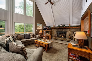 A cozy living room with a stone fireplace, a large window, and a ceiling fan. - Cabin w/ Pond, Hot Tub & Pool Table | Family-Friendly Retreat 3BR, 2BA (Nashville)
