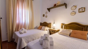 A hotel room with two beds, each with a white bedspread and matching pillows, a wooden headboard, a bedside table with a lamp, and a wall-mounted shelf with decorative items. - Noa de Marchena by Ruralidays (Seville)
