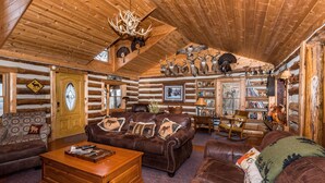 A log cabin interior with a wooden ceiling, a brown leather sofa, a wooden coffee table, and a bookshelf filled with books and decorative items. - Secluded 100-Acre Cabin | Hot Tub, Views & Wildlife 3BR, 1.5BA (Nashville)