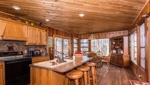 A wooden cabin kitchen with a large island, bar stools, and a dining area with chairs. - Secluded 100-Acre Cabin | Hot Tub, Views & Wildlife 3BR, 1.5BA (Nashville)