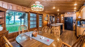A wooden cabin interior with a dining area, kitchen, and large windows overlooking a natural landscape. - Secluded 100-Acre Cabin | Hot Tub, Views & Wildlife 3BR, 1.5BA (Nashville)