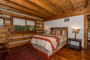 A wooden cabin bedroom with a large bed, two bedside lamps, a wooden dresser, and a window with blinds.