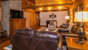 A cozy log cabin interior with a stone fireplace, leather sofas, and a wooden ceiling. - Large Log Cabin | Hot Tub, Game Room & Near Nashville 5BR, 3BA (Nashville)