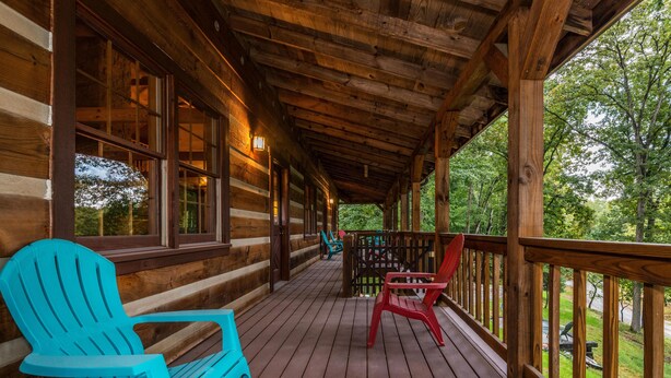 A wooden cabin with a covered porch, featuring two Adirondack chairs and a view of the surrounding forest. - Large Log Cabin | Hot Tub, Game Room & Near Nashville 5BR, 3BA (Nashville)
