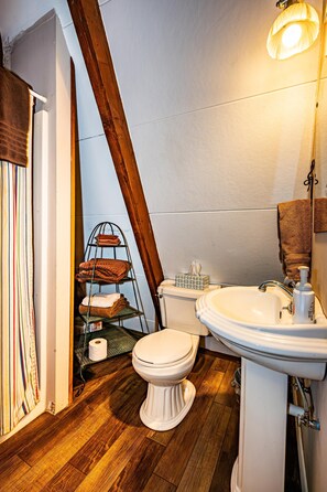 Et badeværelse med trægulv, et hvidt toilet, en vask med sæbedispenser og et håndklædestativ med foldede håndklæder.