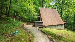Eine Hütte mit geneigtem Dach, umgeben von einem Wald. Ein Kiesweg führt zur Hütte, und eine kleine Terrasse ist auf der rechten Seite sichtbar.