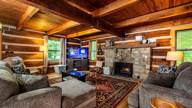 A log cabin interior with a stone fireplace, wooden ceiling beams, and a television in the living area. - Ridge View Log Cabin | Hot Tub, Game Shed & EV Charger 2BR, 2 BA (Nashville)