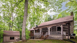 A rustic cabin with a wooden porch and a stone chimney, surrounded by lush greenery. - Ridge View Log Cabin | Hot Tub, Game Shed & EV Charger 2BR, 2 BA (Nashville)