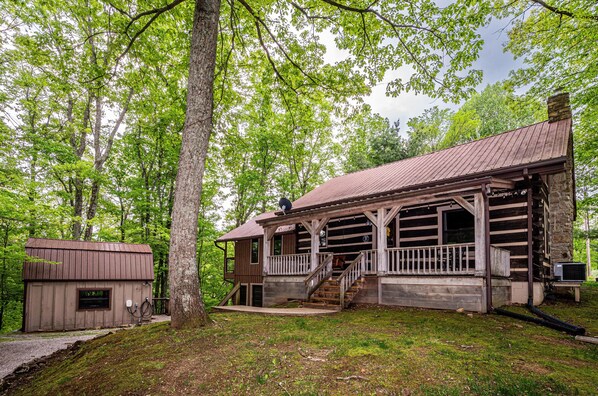 A rustic cabin with a wooden porch and a stone chimney, surrounded by lush greenery. - Ridge View Log Cabin | Hot Tub, Game Shed & EV Charger 2BR, 2 BA (Nashville)