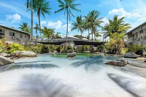 Piscina de resort con agua cristalina, rodeada de palmeras y rocas, con edificios al fondo.