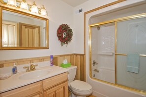 A bathroom with a large mirror, a sink with a gold faucet, a toilet, and a bathtub with a glass door. - Log Cabin Near Nashville | Hot Tub, Screened Porch & Game Loft2BR, 2BA (Nashville)