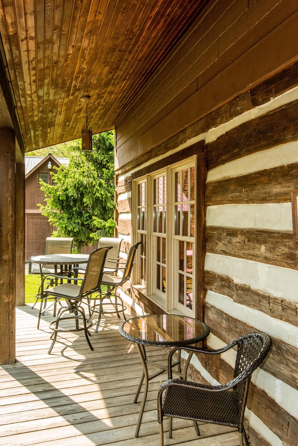 A wooden deck with a glass table and chairs, a wooden cabin with a window, and a tree in the background.