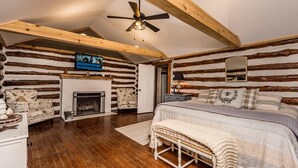 Un dormitorio de estilo cabaña de troncos con chimenea, vigas de madera, una cama, un banco y un ventilador de techo.