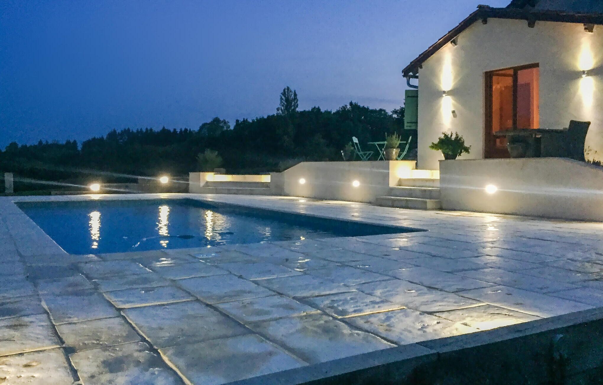 Piscina com deck revestido de azulejos, rodeada por um muro baixo e um edifício com iluminação externa.