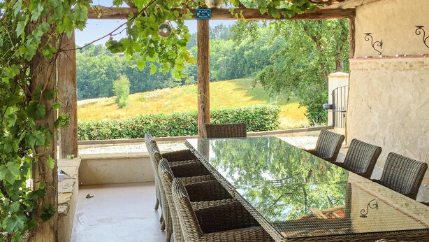 Un espacio de comedor al aire libre techado, con mesa de vidrio y sillas de mimbre, con vistas a un paisaje verde.