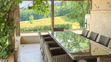 Un espace extérieur abrité, aménagé pour les repas, avec une table en verre et des chaises en osier, offrant une vue sur un paysage verdoyant.