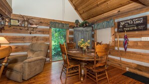 Interior de una cabaña de madera con una mesa de comedor, sillas, un sillón reclinable y un letrero que anuncia alojamientos para cazadores.