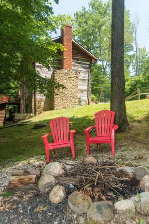 A rustic cabin with a stone exterior and a chimney, surrounded by trees and a fire pit with logs.