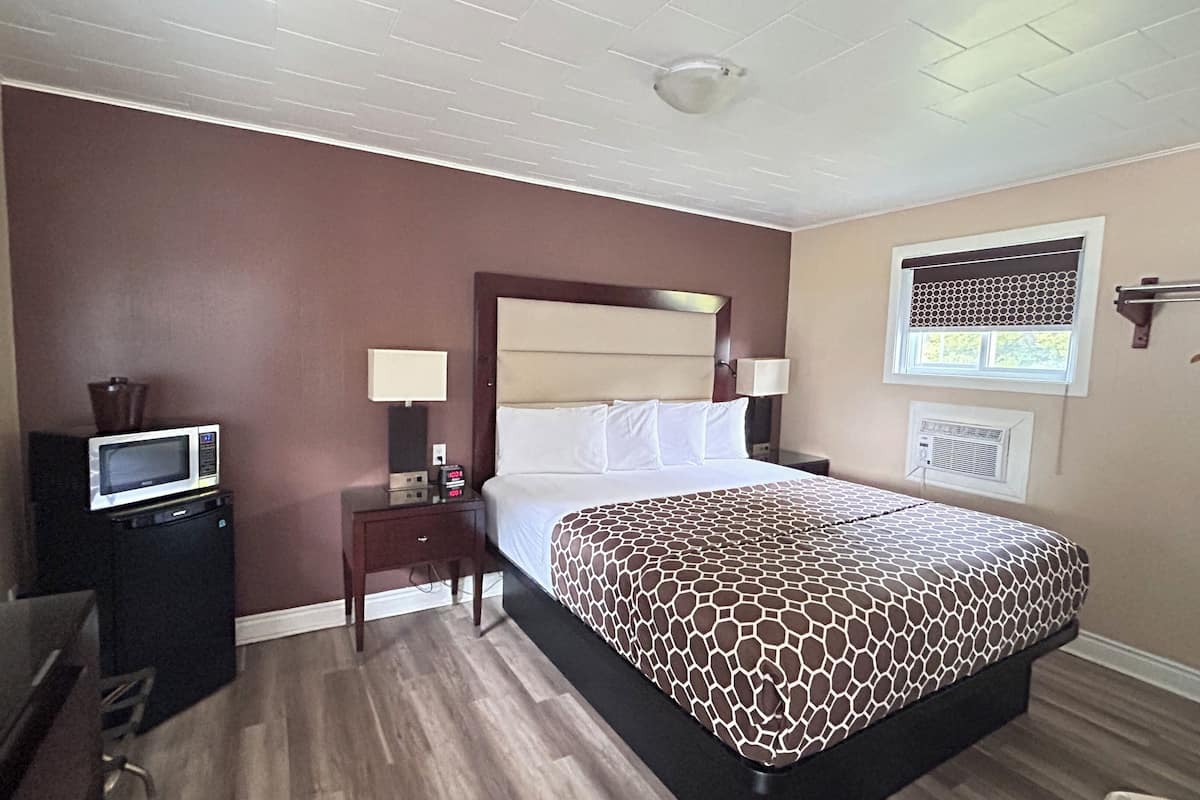 A hotel room with a bed, a desk with a microwave, a nightstand, and a window with blinds.