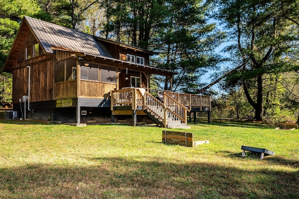 Rustic cabin exterior with a metal roof, screened porch, wooden deck, and grassy yard featuring cornhole boards.