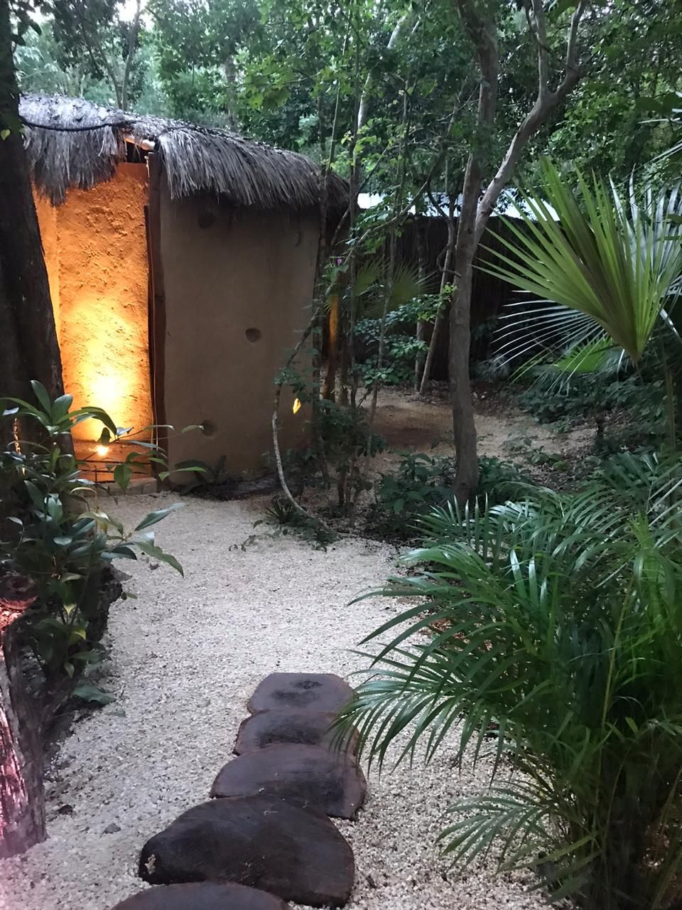 Un sendero conduce a una cabaña rústica con techo de paja, rodeada de follaje y suelo arenoso.
