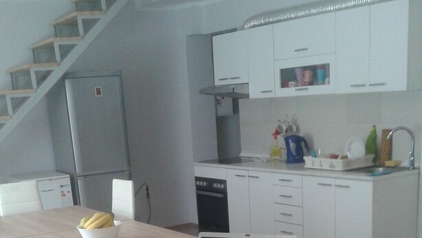 A modern kitchen with white cabinets, a dishwasher, and a sink. There's a wooden dining table with chairs and a laptop on it. - Hostel A&A (Novi Sad)