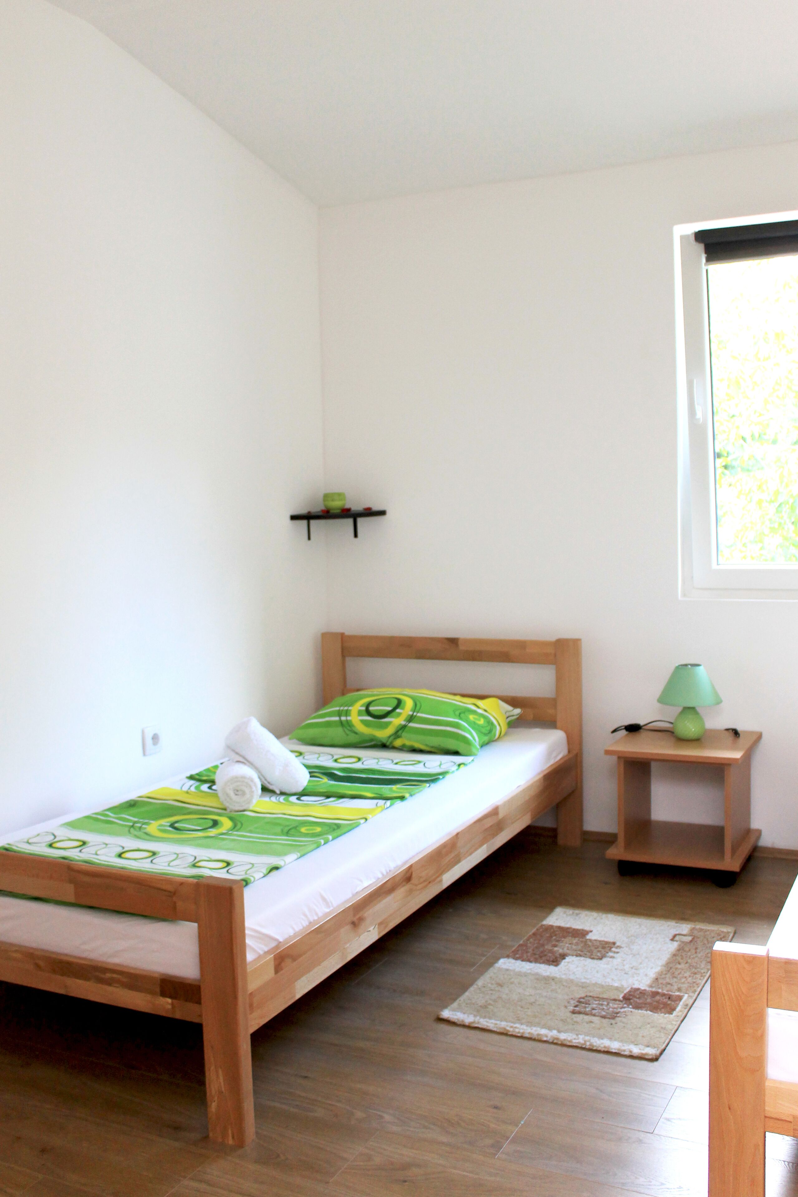 A single bed with a wooden frame, a green and white patterned bedspread, and a small bedside table with a green lamp.