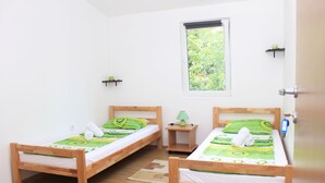 A room with two wooden beds, a window with a view of greenery, a small nightstand with a lamp, and a wall-mounted shelf with a green apple. - Hostel A&A (Novi Sad)