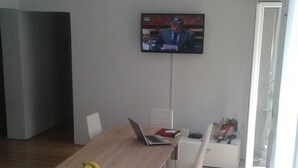 A modern dining area with a long wooden table, white chairs, a laptop, and a bowl of fruit. A television is mounted on the wall. - Hostel A&A (Novi Sad)