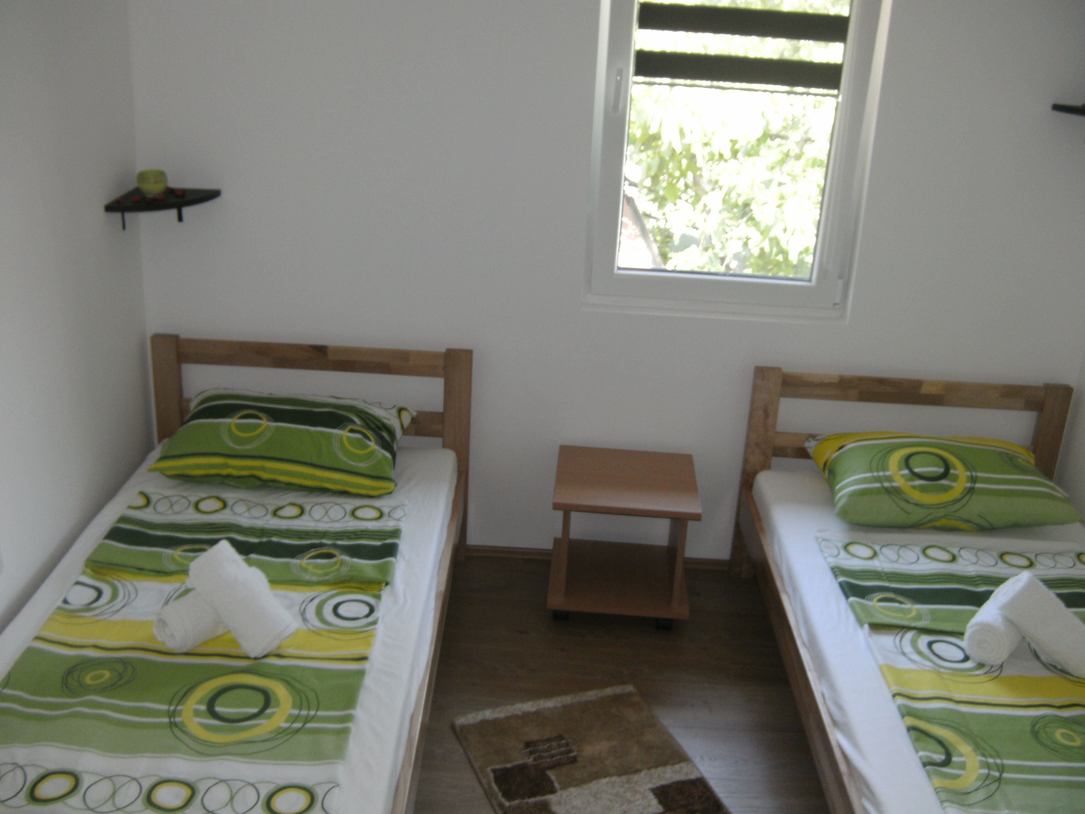 A room with two single beds, a small wooden nightstand, and a window with blinds.