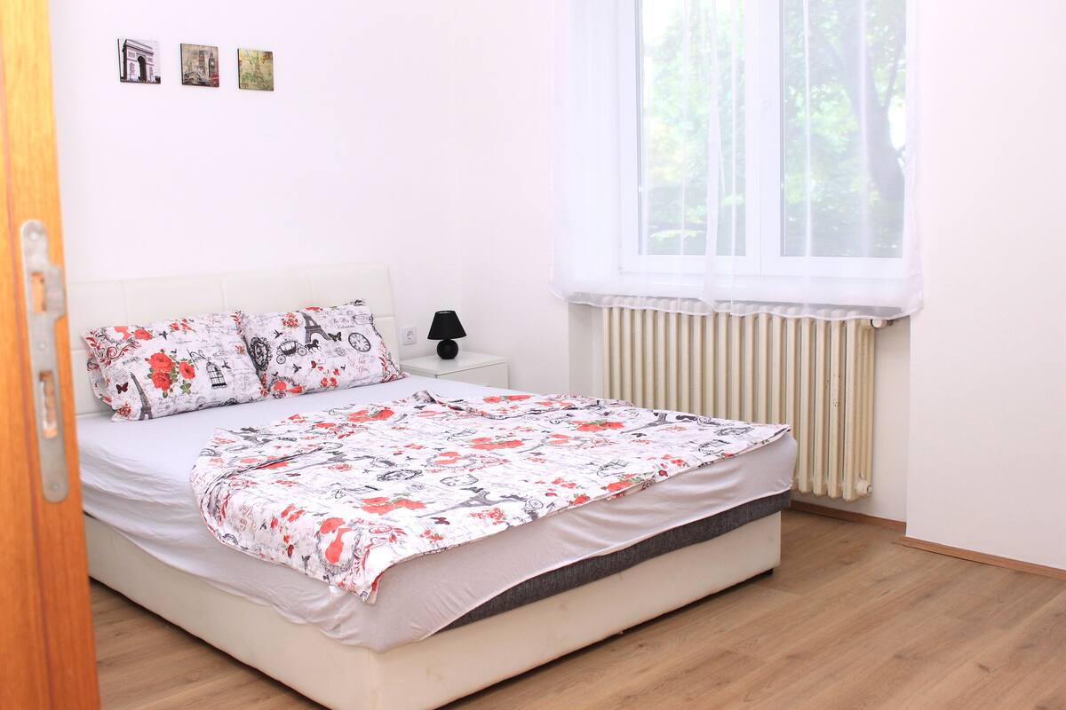 A neatly made bed with floral bedding, a bedside table with a lamp, and a window with sheer curtains.