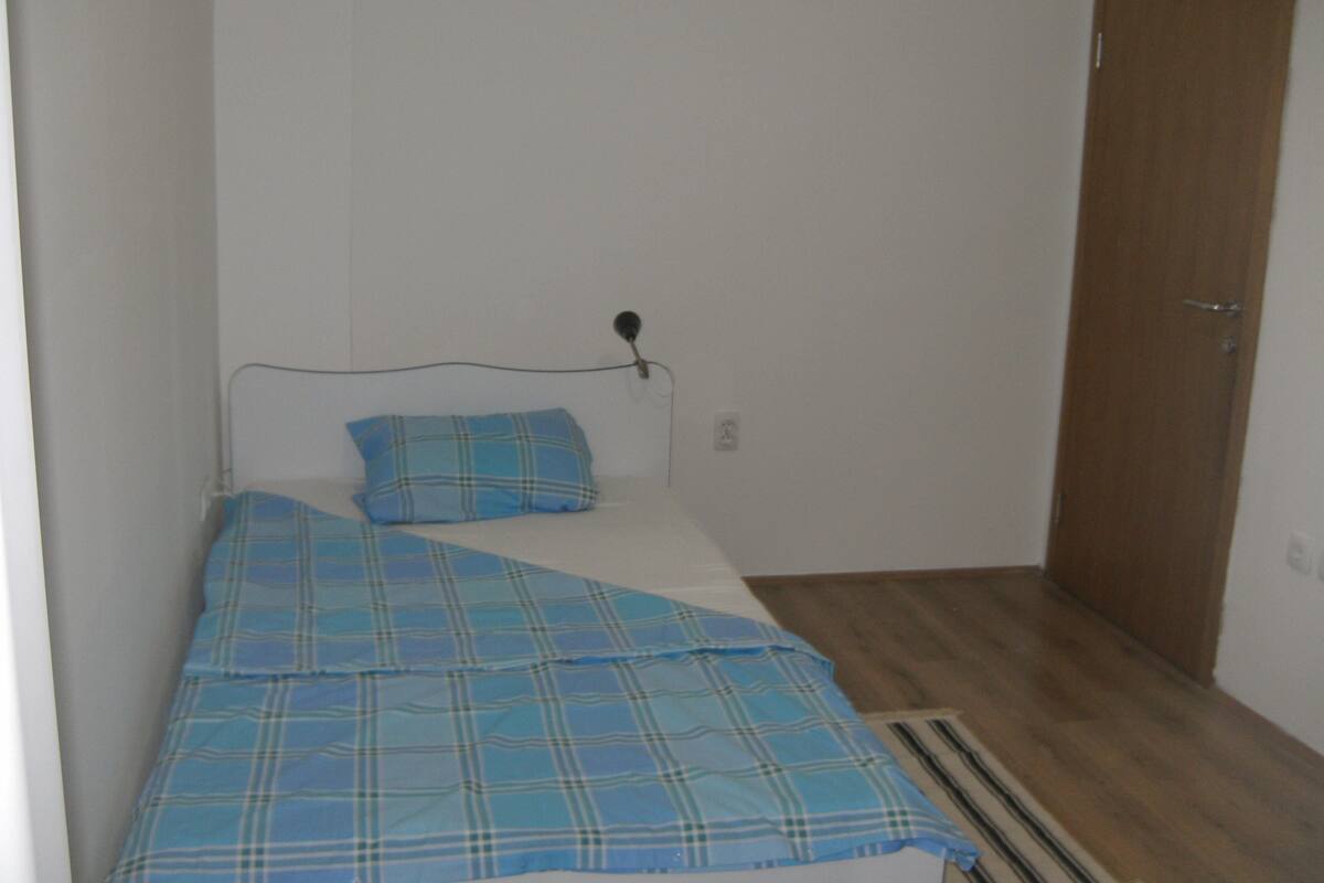 A small, single-bed room with a wooden floor, a metal bed frame, and a blue plaid bedspread.