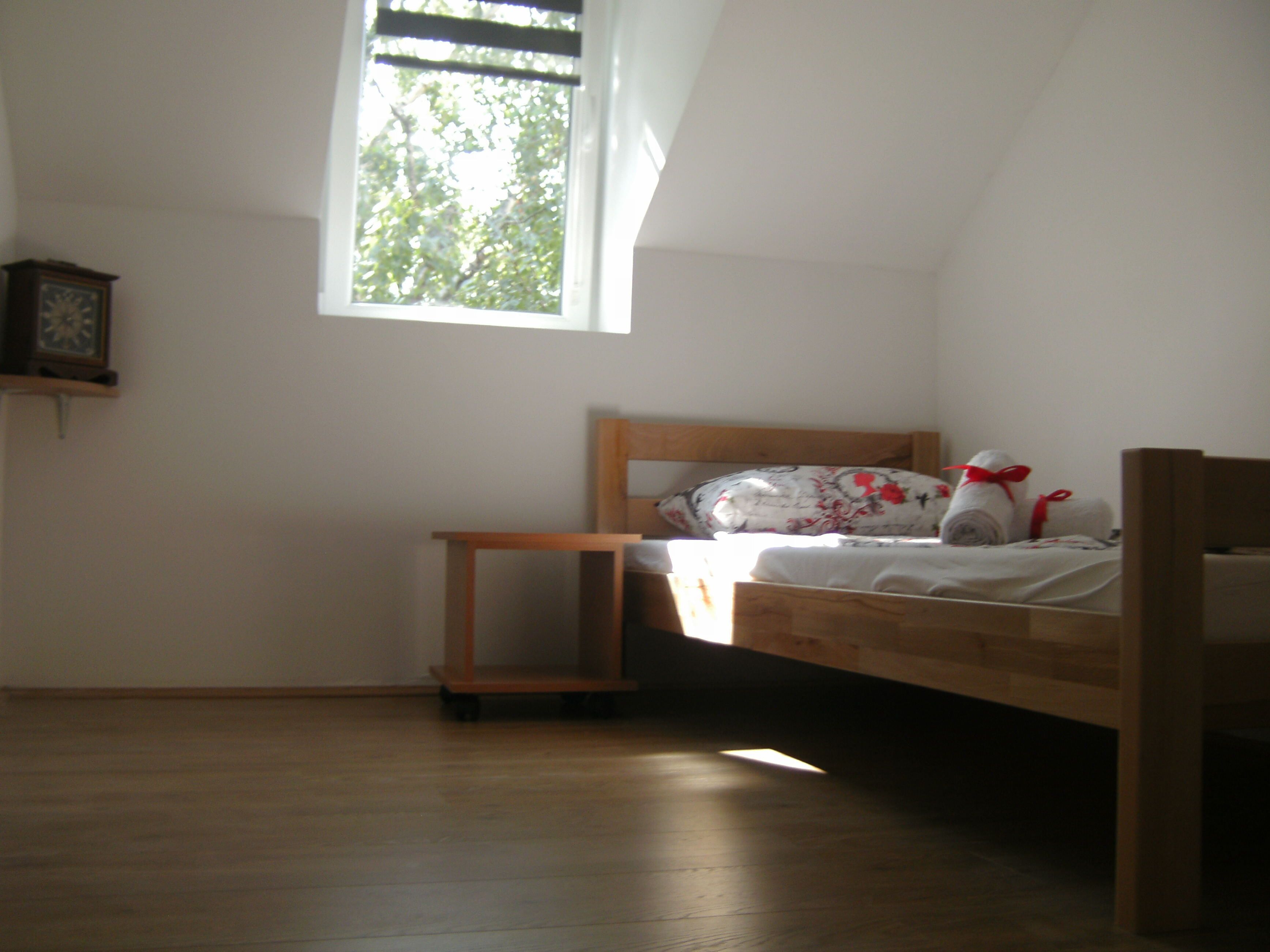 A small, single-bed room with a wooden headboard, a wooden nightstand, a small table, and a window with a view of trees.