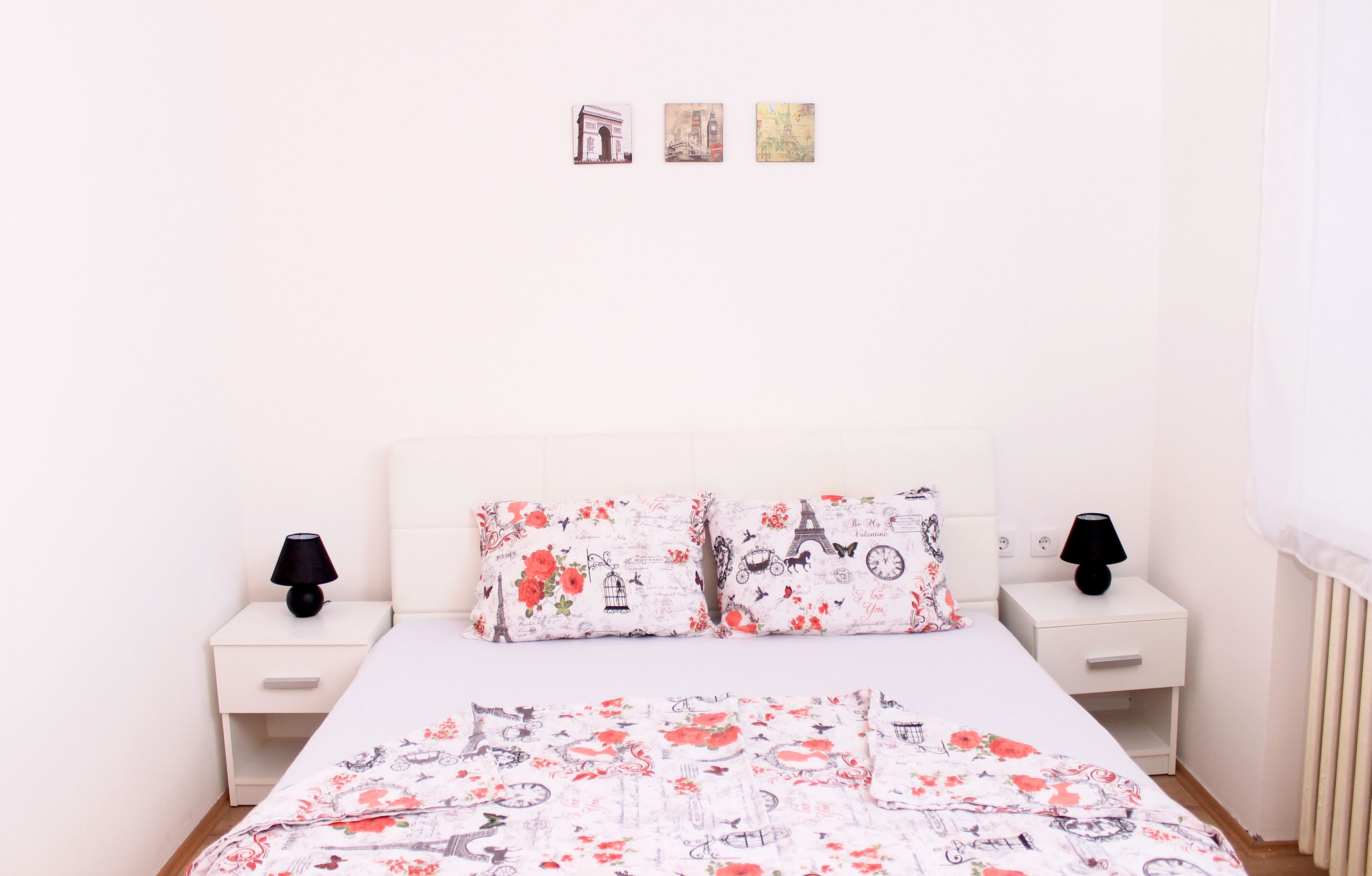 A bedroom with a white bed, floral bedding, two nightstands with lamps, and a plain wall with three small pictures.