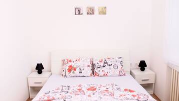 A bedroom with a white bed, floral bedding, two nightstands with lamps, and a plain wall with three small pictures.