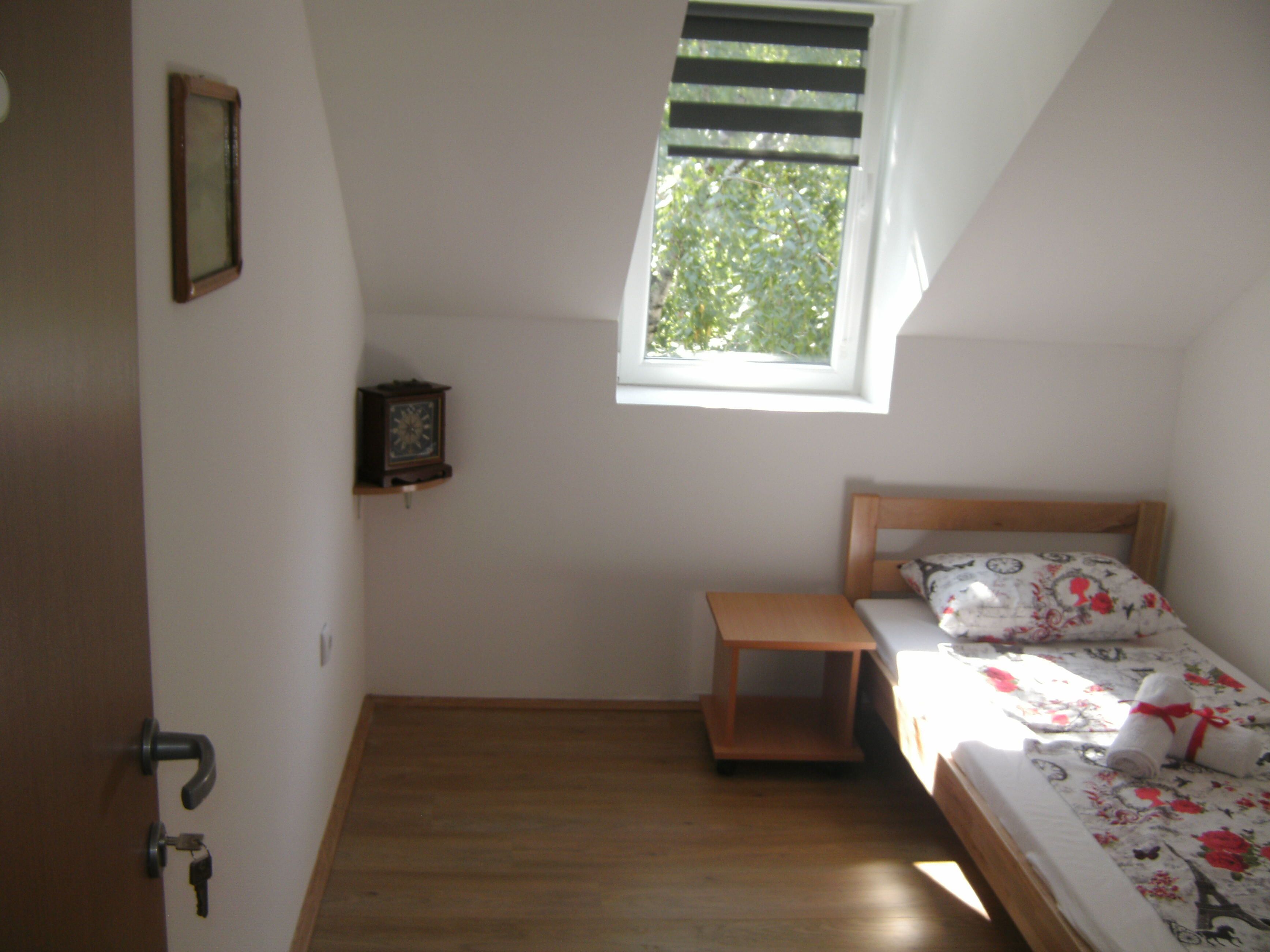 A small, single-bed room with a wooden headboard, a bedside table, a small shelf with a clock, and a window with blinds.