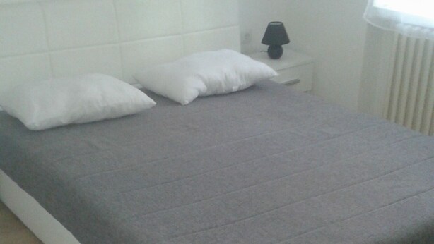 A bedroom with a bed, white headboard, bedside table with a lamp, and a window with blinds. - Hostel A&A (Novi Sad)