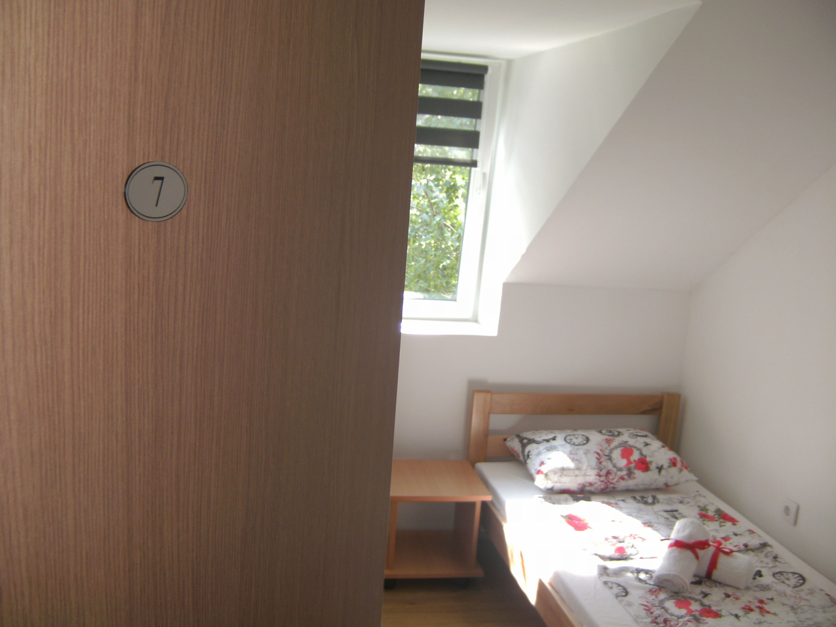 A small, single-bed room with a wooden door marked with the number 7, a window with blinds, and a bedside table.