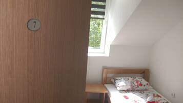 A small, single-bed room with a wooden door marked with the number 7, a window with blinds, and a bedside table.