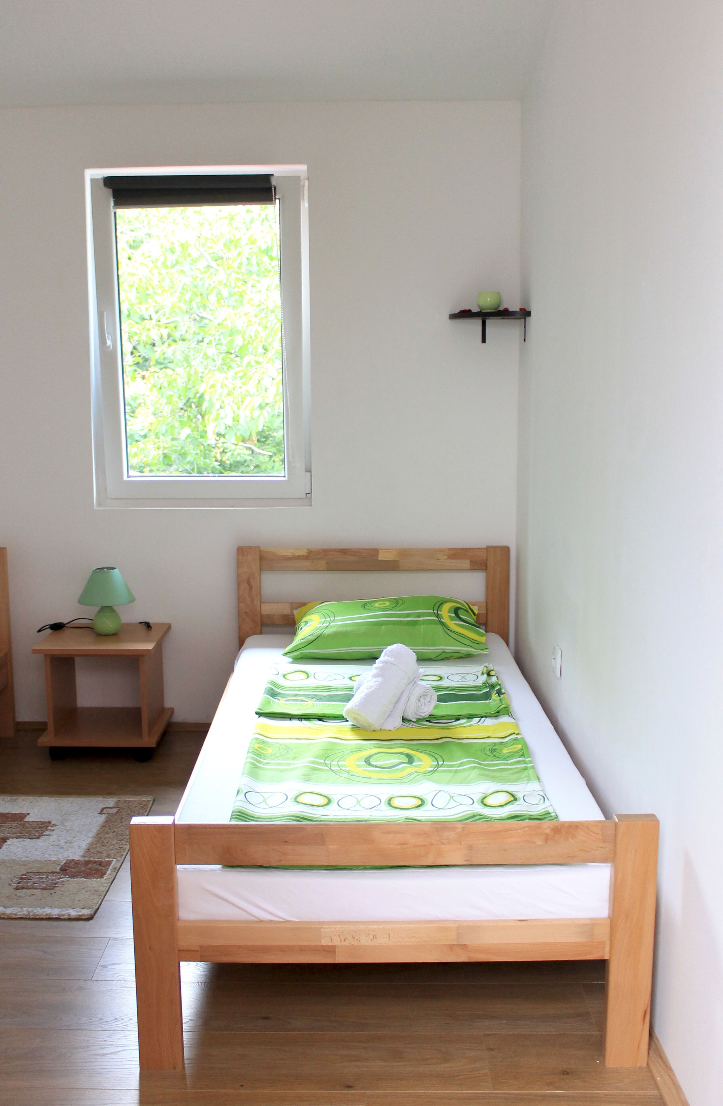 Una cama pequeña y bien hecha con una colcha de diseño blanco y verde, un cabecero de madera y una mesita de noche con una lámpara verde.