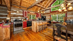 Een rustiek interieur van een chalet met een houten plafond, een centraal eiland met krukken, een grote eettafel en een keuken met rode kasten en een fornuis.