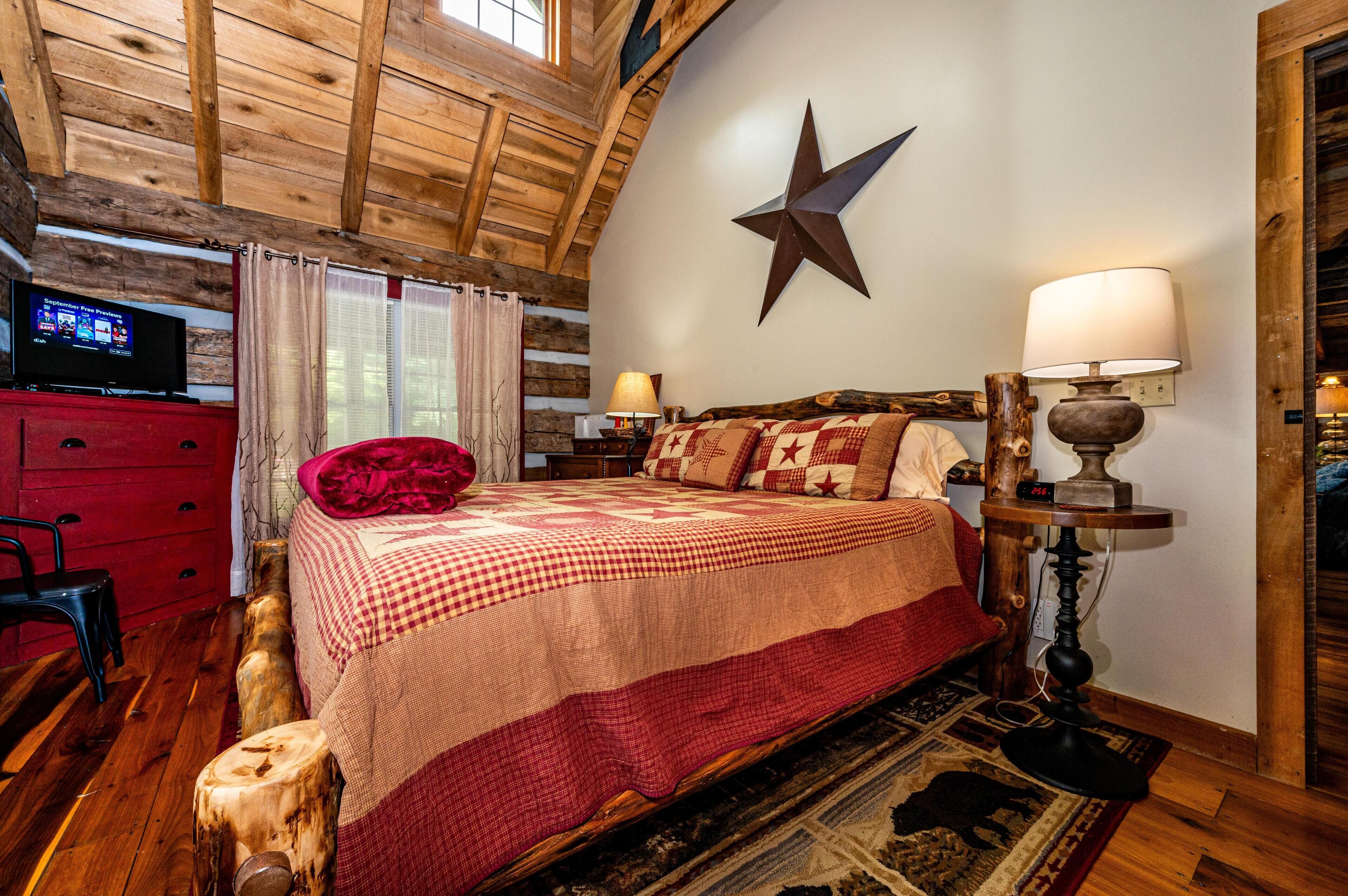 A cozy room with a wooden ceiling, a star decoration on the wall, a bed with a checkered blanket, a red chair, a wooden stool, and a TV on a red cabinet.