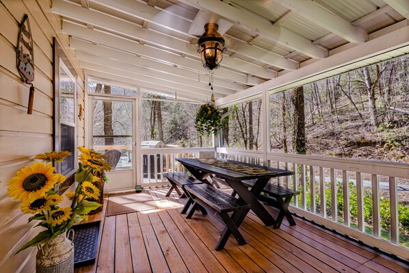 Una terraza de madera con una mesa de picnic y bancos, rodeada de barandillas blancas y grandes ventanales que ofrecen vista al bosque.