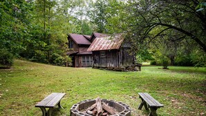 A rustic cabin surrounded by a lush forest, with a fire pit and two picnic tables in the foreground. - 4 BR, 2 BA Log Cabin, charming mix of old and new, close to village of Story (Nashville)