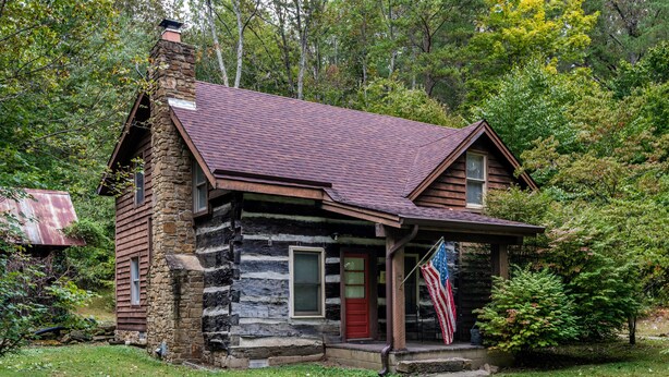 A rustic log cabin with a stone chimney, a red door, and an American flag on the porch. - 4 BR, 2 BA Log Cabin, charming mix of old and new, close to village of Story (Nashville)