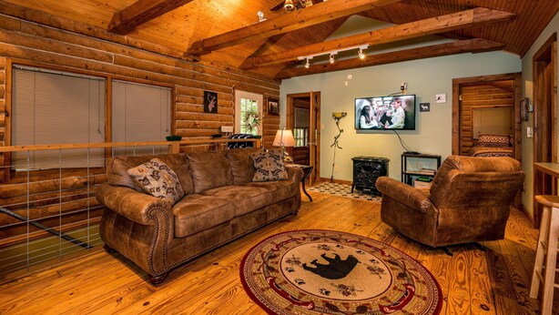 Ein Holzhaus-Interieur mit Holzbalkendecke, einem Ledersofa, einem passenden Sessel, einem Fernseher und einem Kamin.