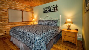 Ein Schlafzimmer in einer Blockhütte mit einem Bett, zwei Nachttischen mit Lampen, einem Wandbild von Bergen und Tieren sowie einem Deckenventilator.