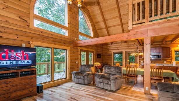 A wooden cabin interior with a television displaying "dish cinema," a dining area, and a kitchen. - View Cabin w/ Hot Tub, Pond & Pool Table | Secluded Retreat 3BR, 2BA (Nashville)