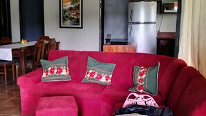 Ein Wohnzimmer mit rotem Sofa, passendem Hocker und einem Essbereich mit Tisch und Stühlen. Im Hintergrund ist eine Küche mit Schränken und Geräten zu sehen.