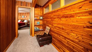 Interior acolhedor de uma cabana com paredes de madeira, um pequeno espaço de estar com um assento almofadado, e uma cama visível ao fundo.